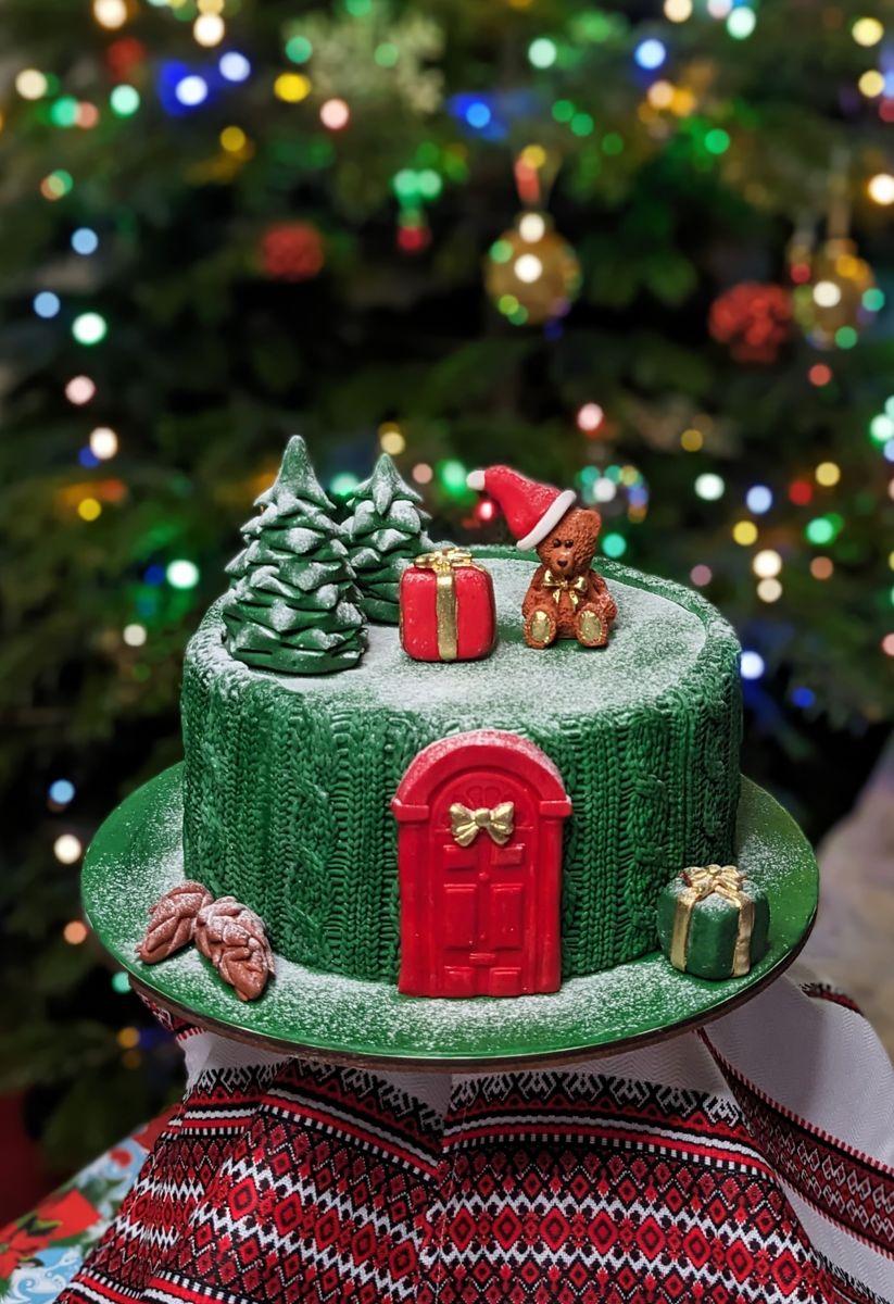 Christmas cake in green knitted-effect fondant with a red door, snowy trees and a teddy bear on top