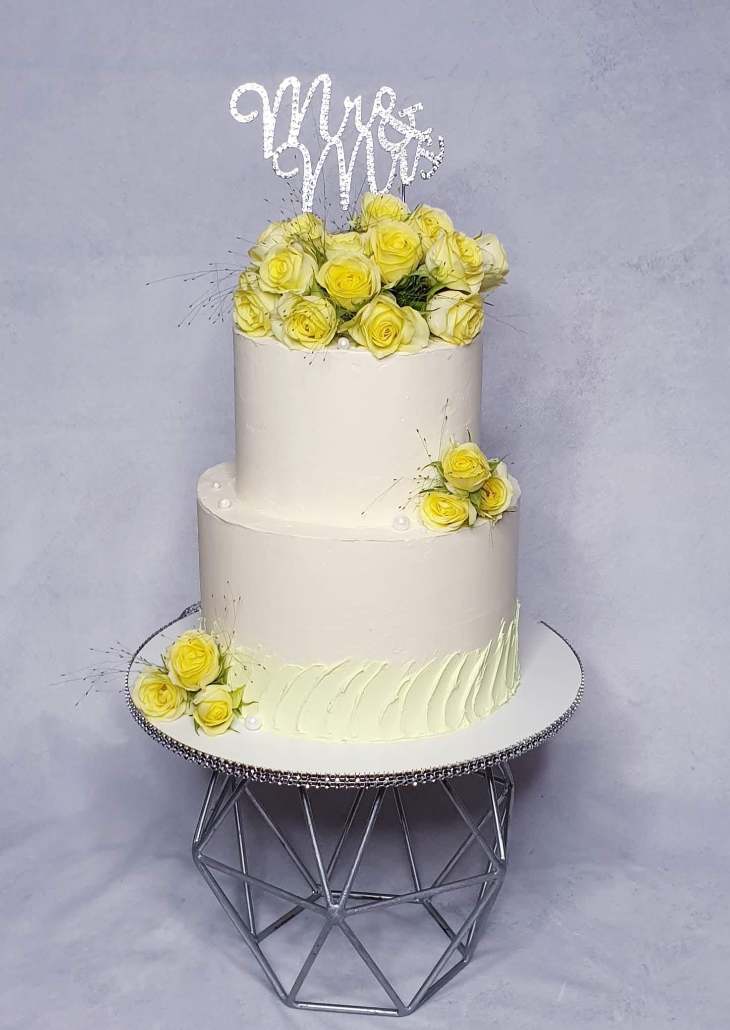 Two-tier white wedding cake with fresh yellow roses, decorative piped detail, and silver 'Mr & Mrs' topper on geometric stand