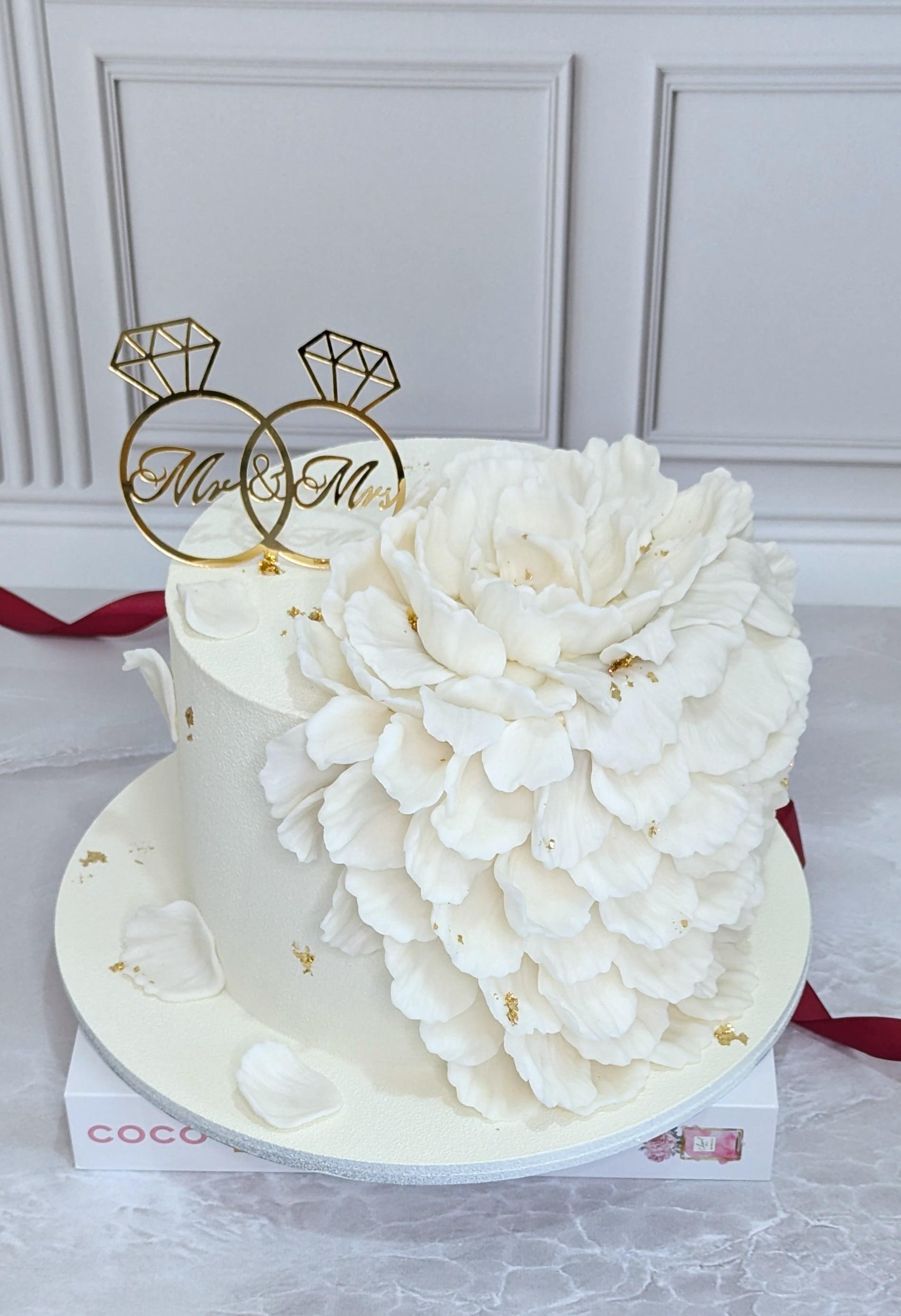 Two-tier white wedding cake with cascading sugar flower petals and gold accents, topped with interlocking diamond ring topper