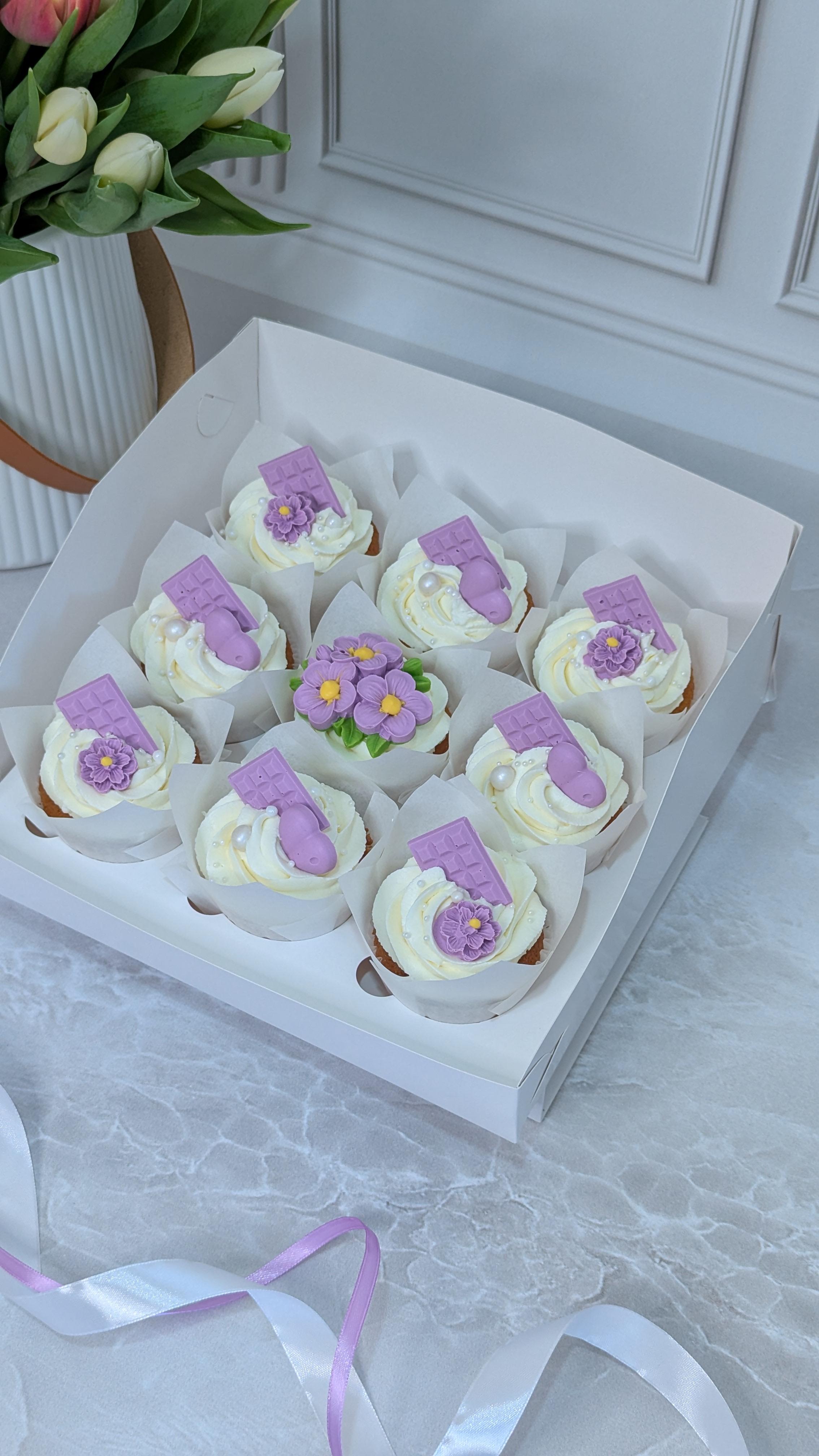 Nine vanilla cupcakes with white buttercream, purple wafer toppers and small purple fondant flowers in a white box