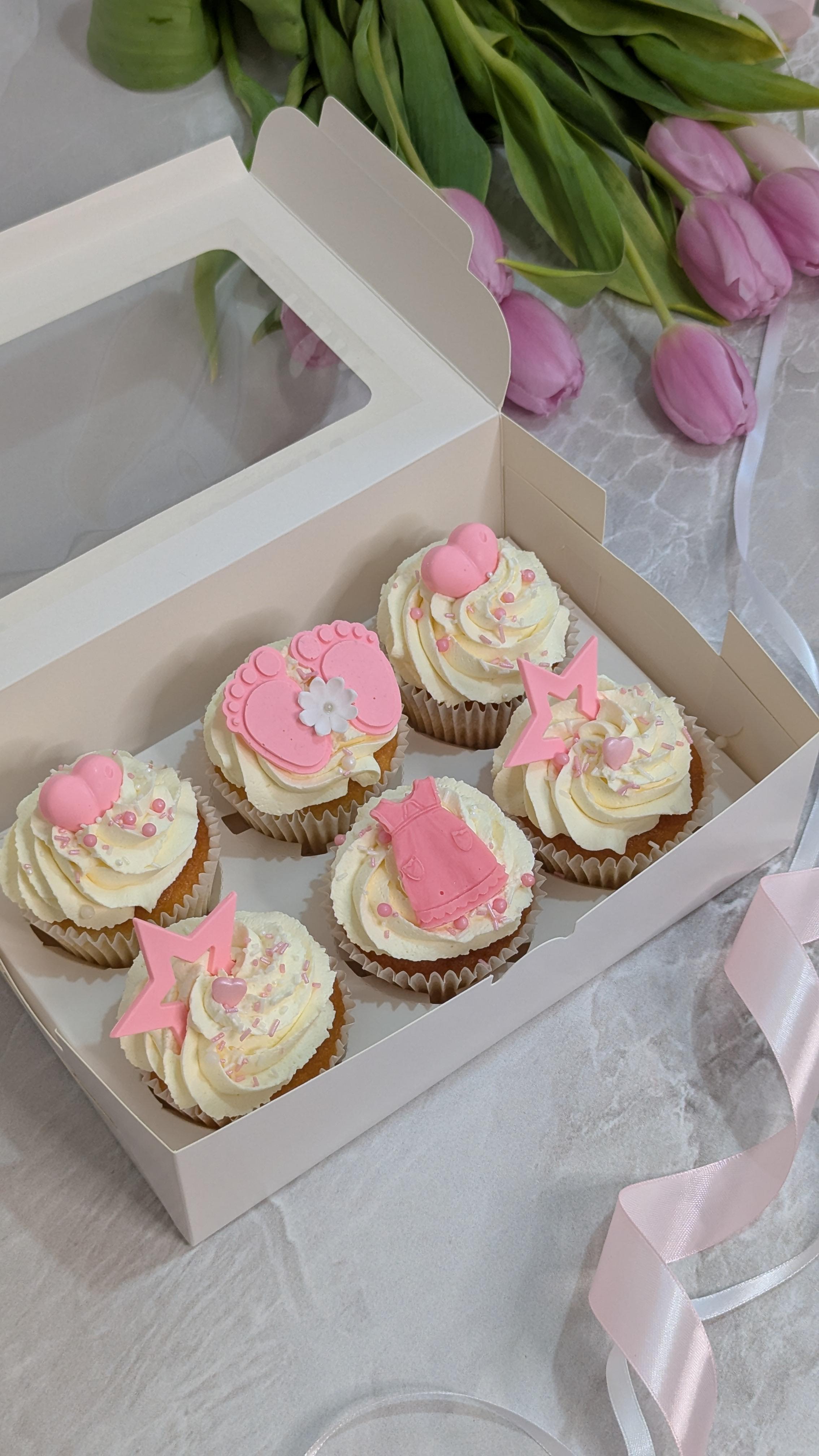 Six cream-frosted cupcakes with pink fondant decorations including hearts, scalloped shapes, flowers and stars