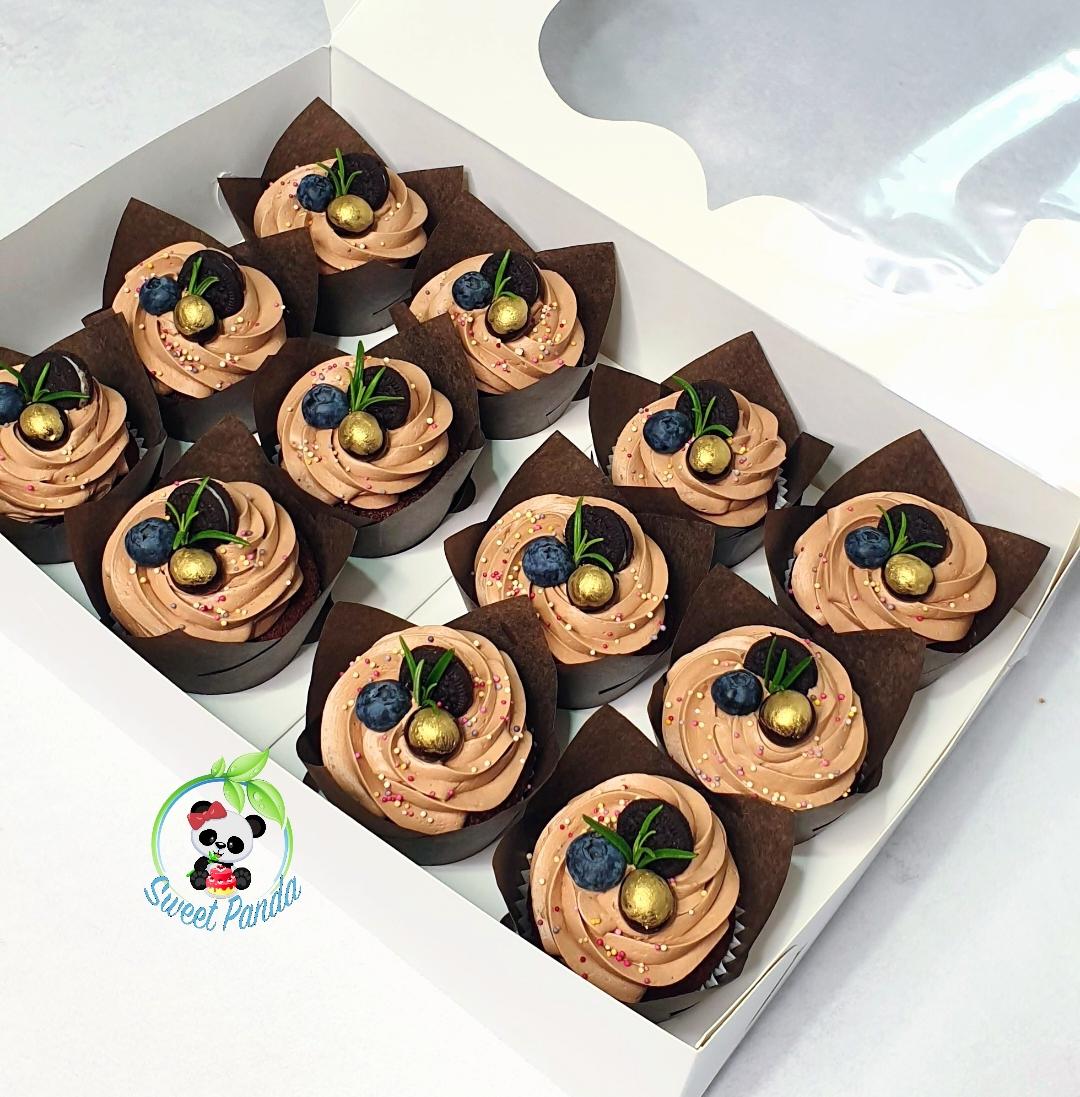 Twelve chocolate cupcakes with swirled frosting, topped with blueberries, gold pearls and rosemary sprigs, in brown cases