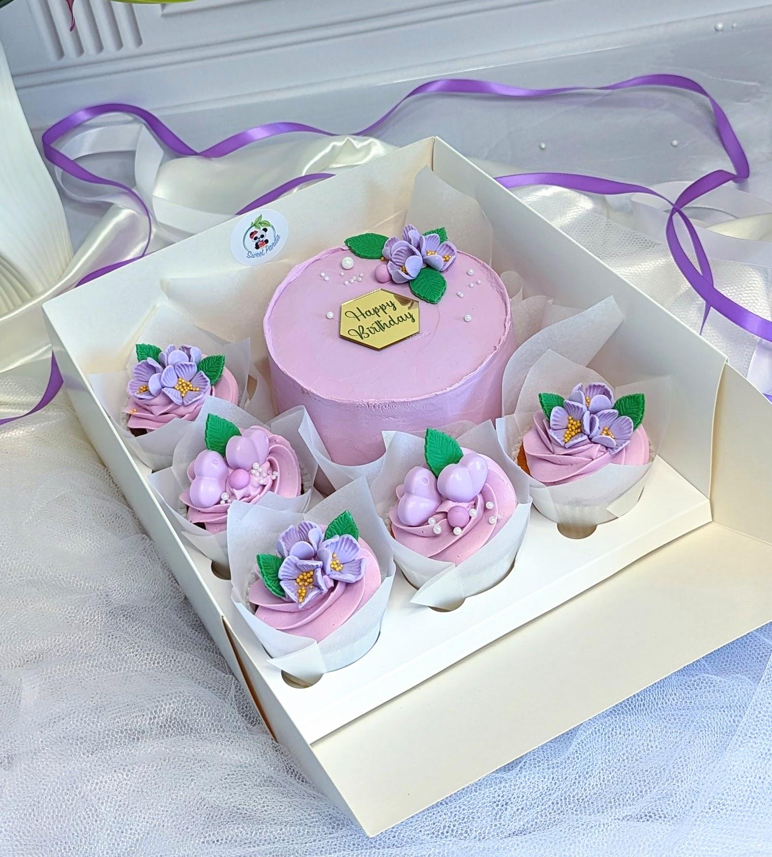 Bento box with pink round cake and four pink cupcakes, decorated with purple and lavender sugar flowers and green leaves