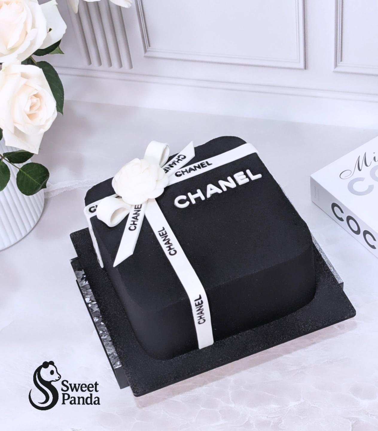 Black square fondant cake with white CHANEL-branded ribbon and white camellia flower on grey cake board