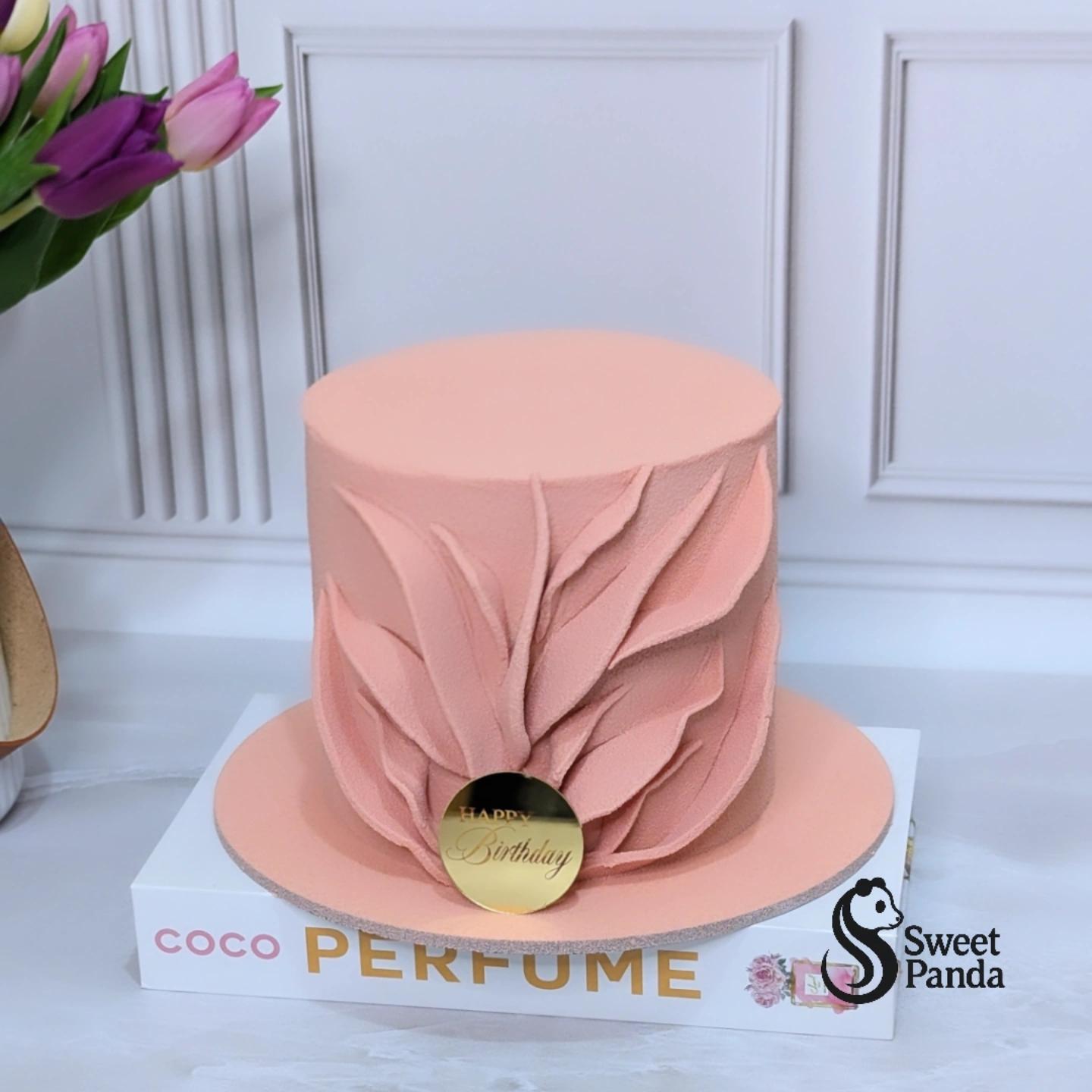 Single-tier blush pink cake with textured petal design and gold circular 'Happy Birthday' topper on white cake board