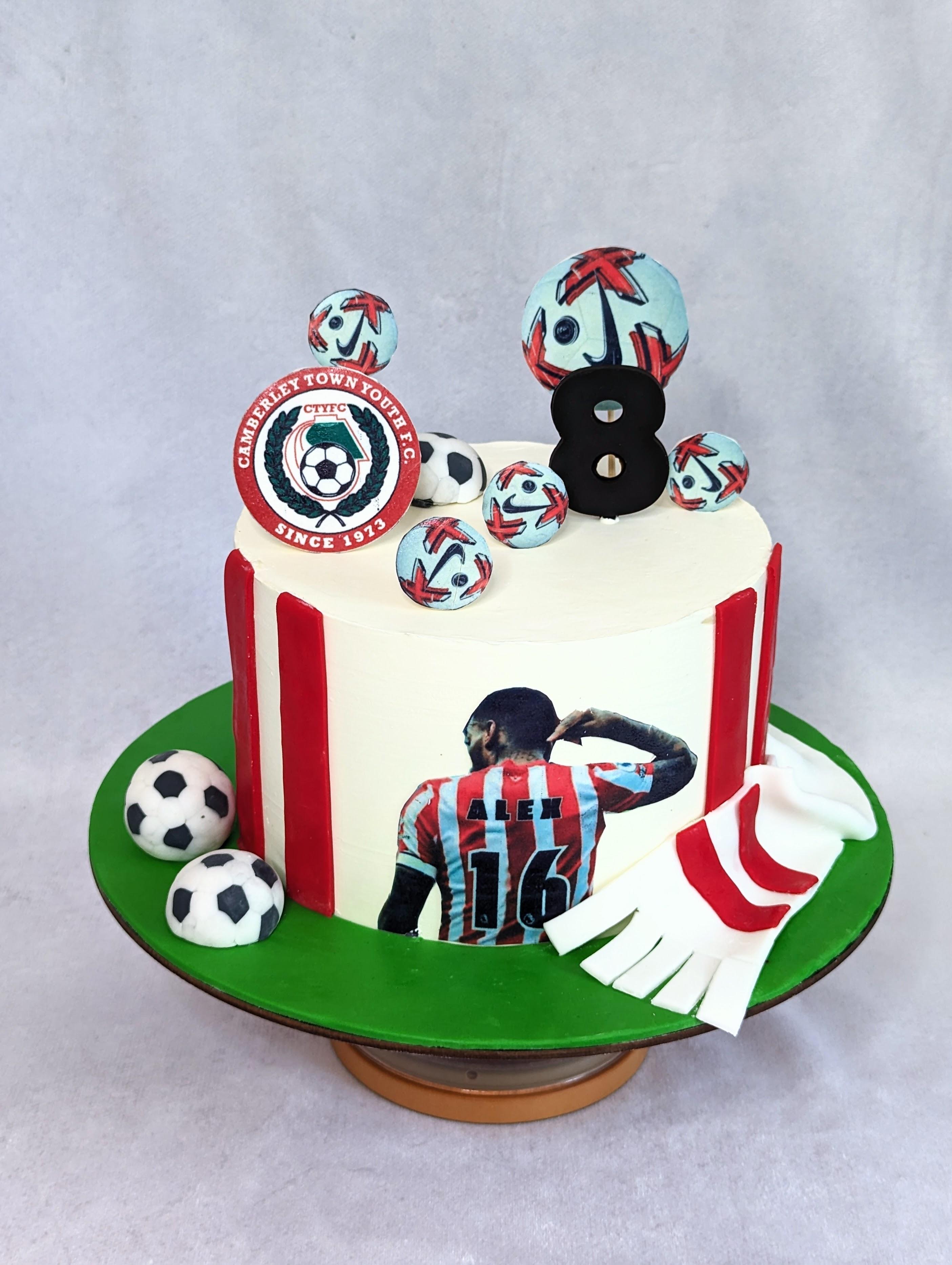 Football-themed cake with red vertical stripes, cream fondant, green grass base, soccer balls