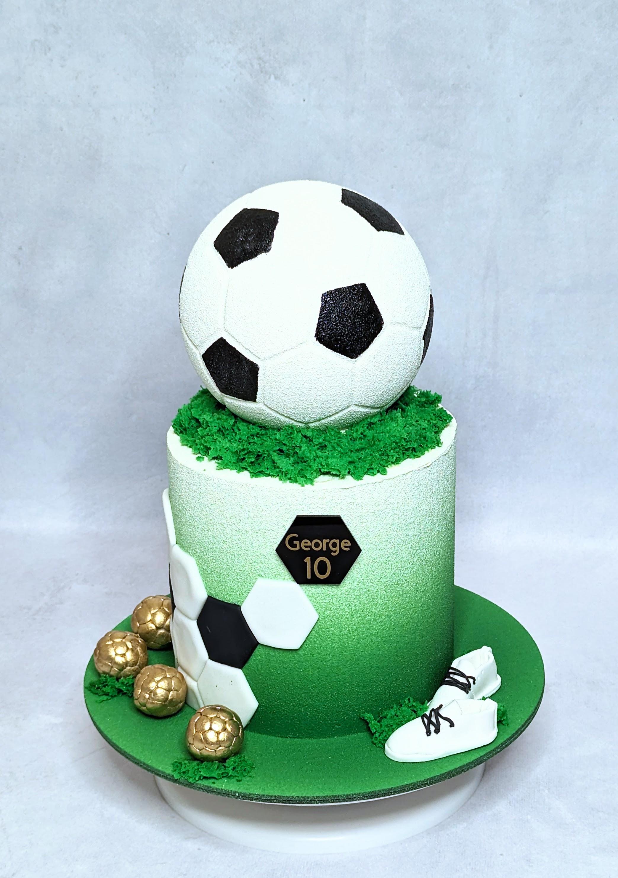 Two-tier football-themed birthday cake with white soccer ball topper, green fondant base, gold chocolate balls