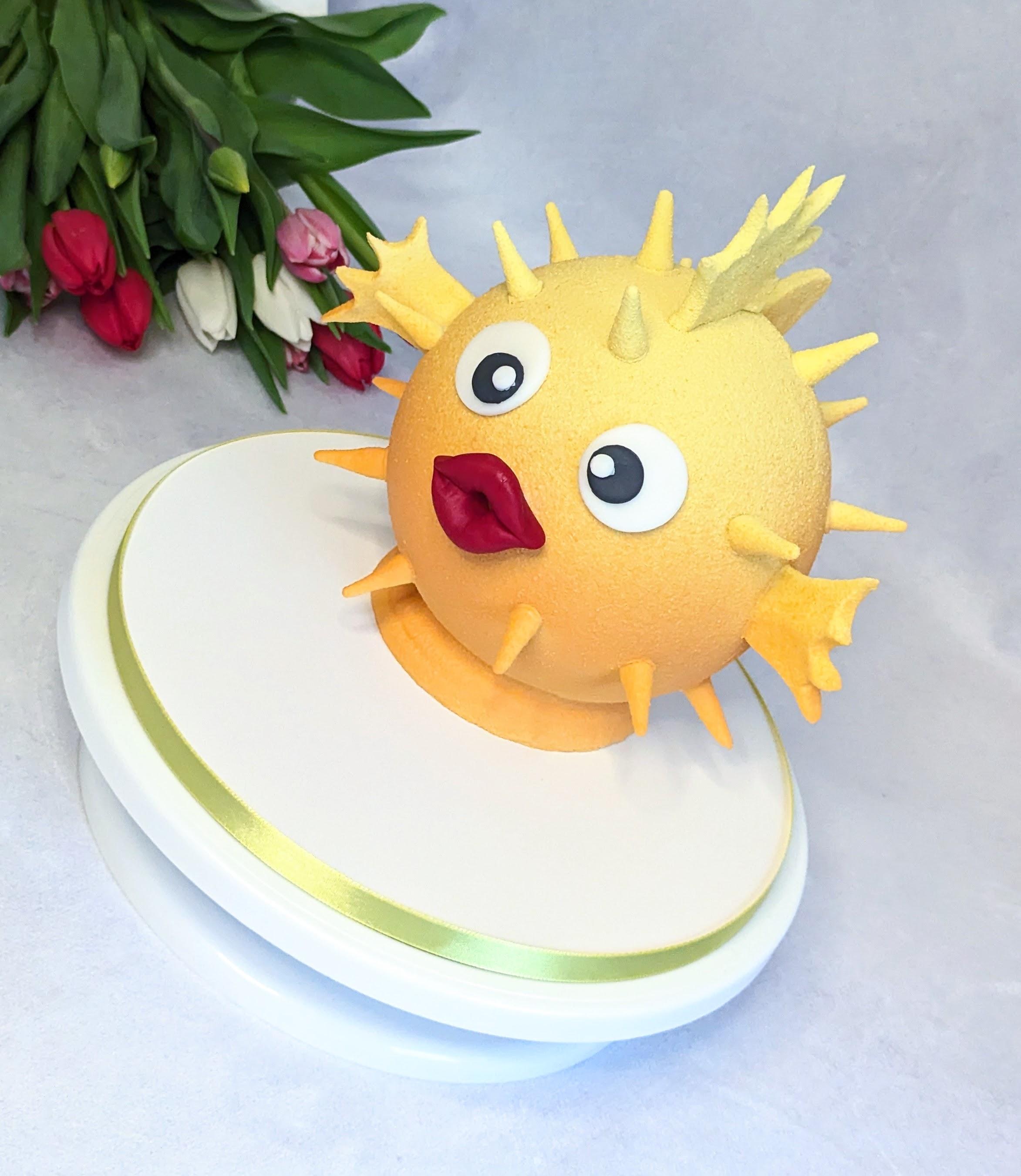 White cake with gradient green and blue border topped with a yellow spiky fondant creature with large eyes and open mouth