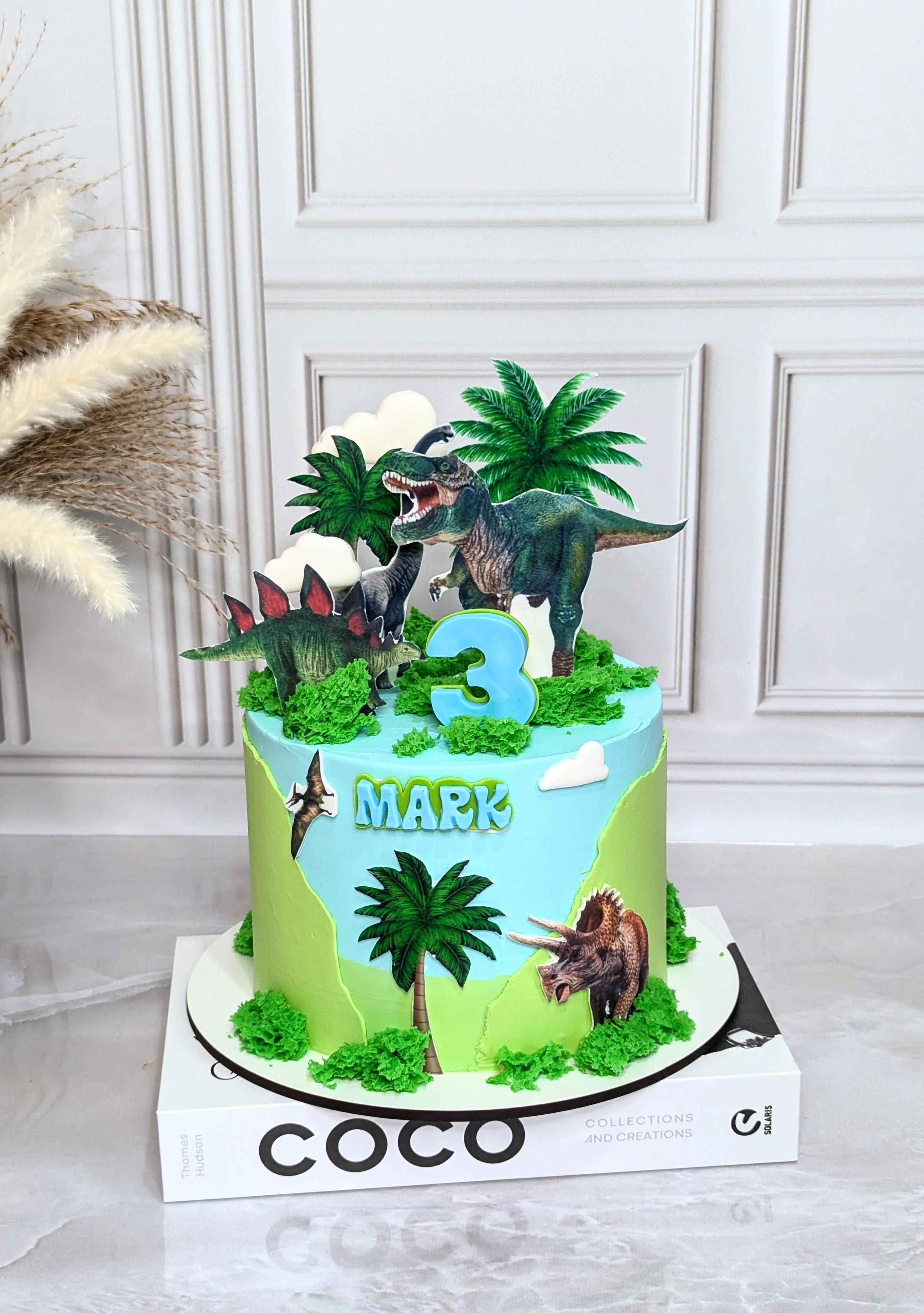 Two-tier dinosaur birthday cake with lime and blue fondant, featuring printed dinosaur images, palm trees, green moss texture