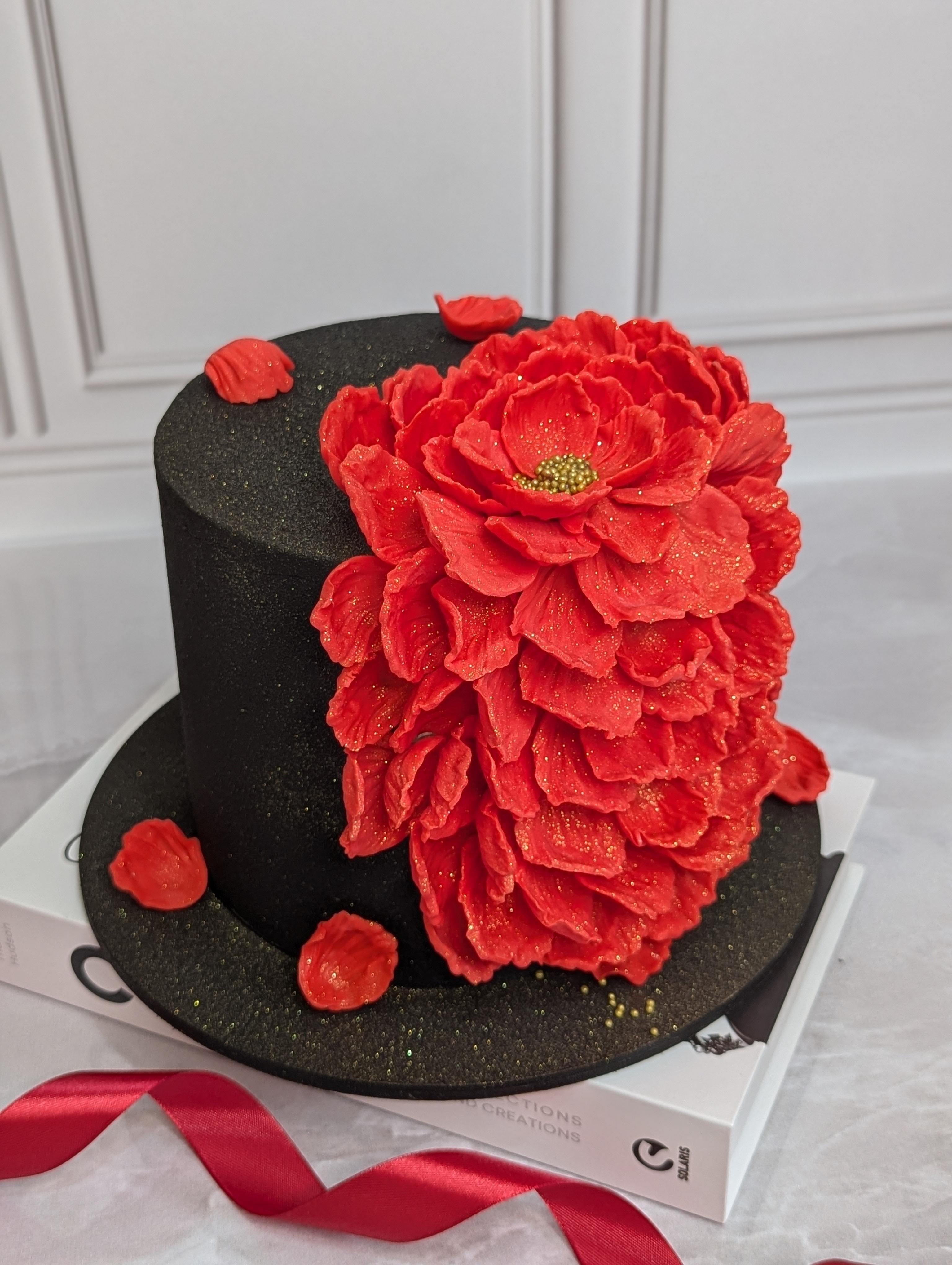 Two-tier black cake with large red sugar peony flower and small red petals, dusted with gold sparkle