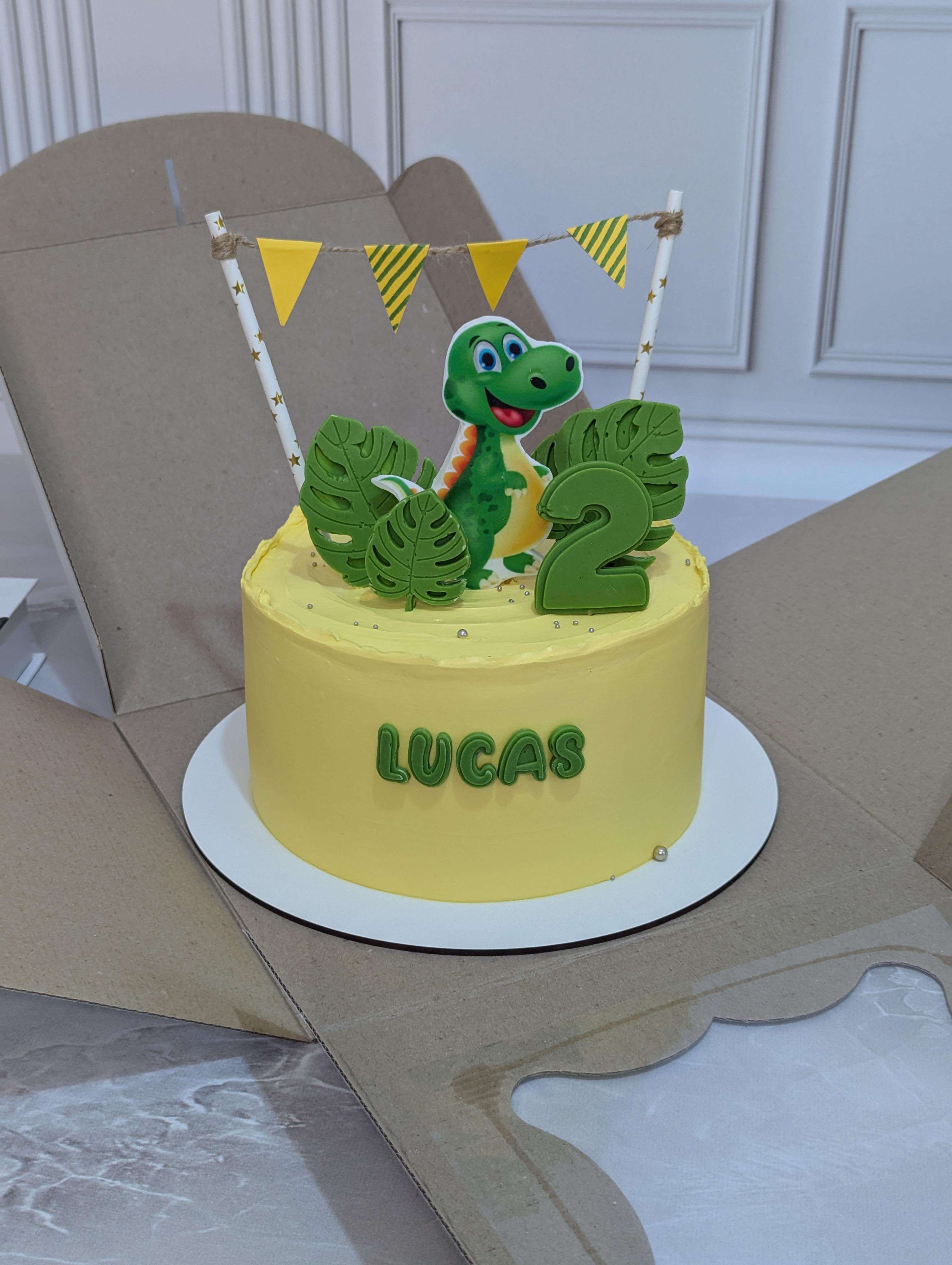 Single-tier yellow cake with green dinosaur figurine, number 2 topper, monstera leaf decorations and yellow bunting