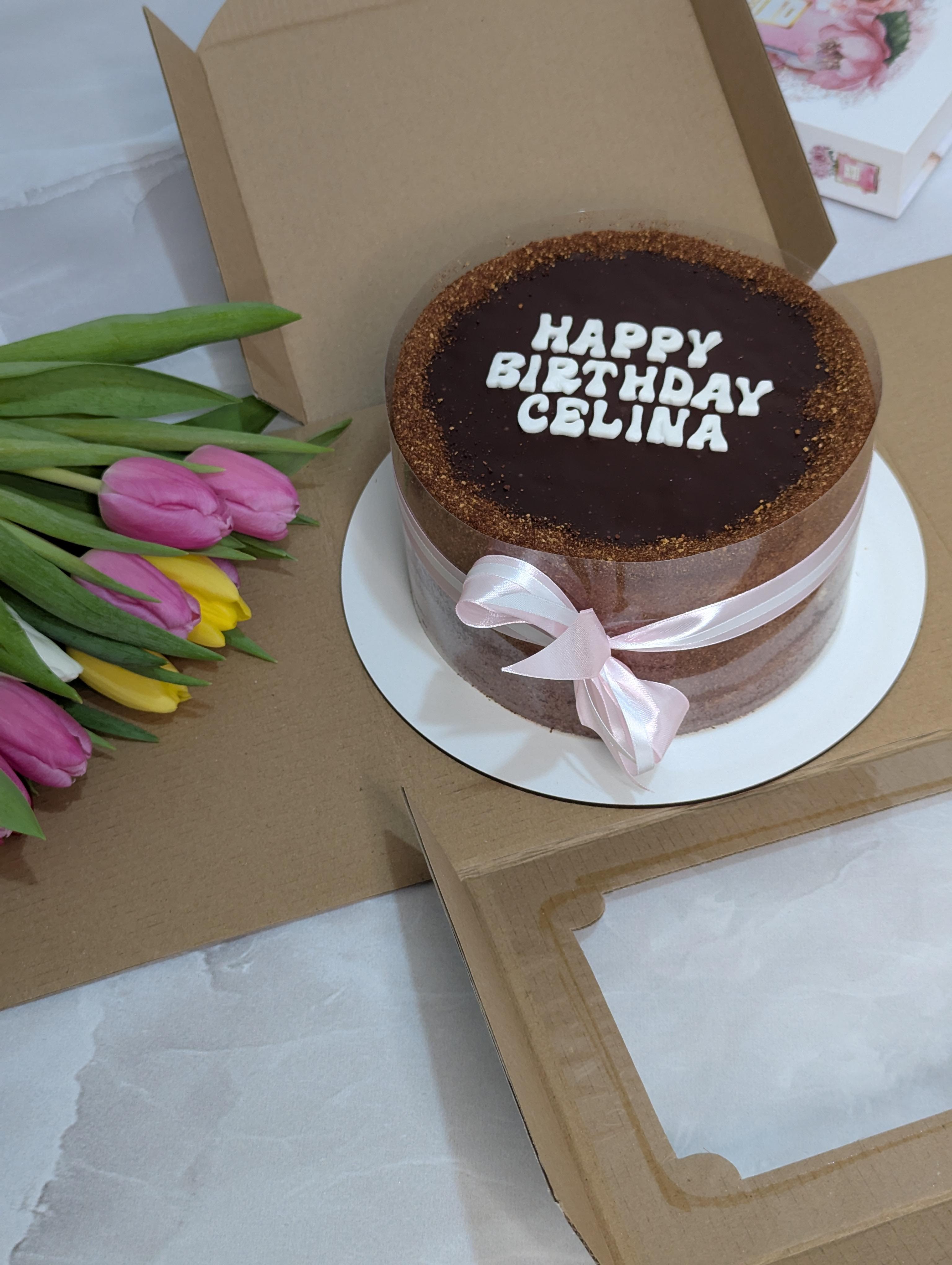 Two-tier chocolate cake with dark ganache top, gold sparkly edge, white piped message and pink ribbon bow