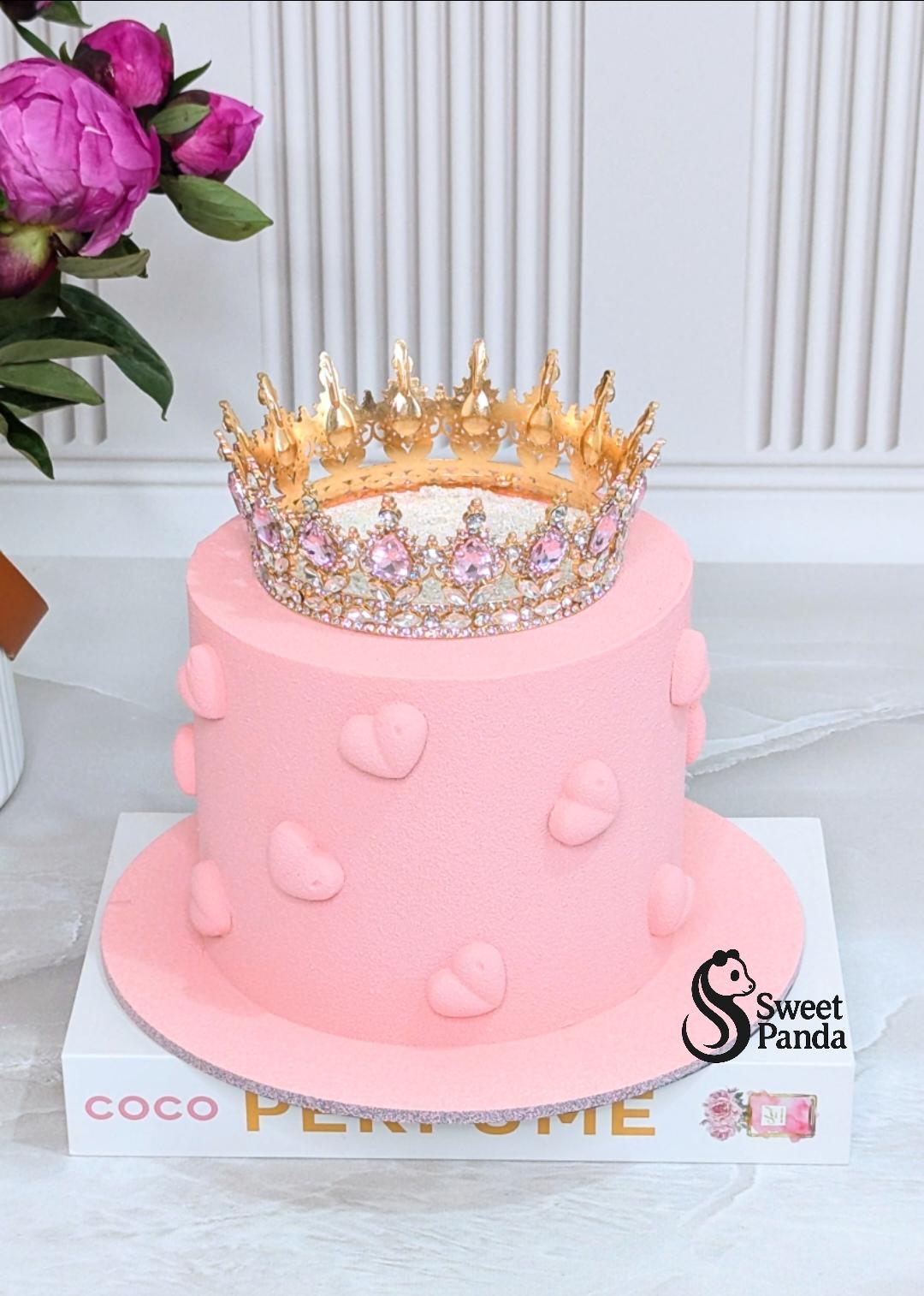 Two-tier pink fondant cake with heart decorations and ornate gold crown topper, decorated with magenta peonies