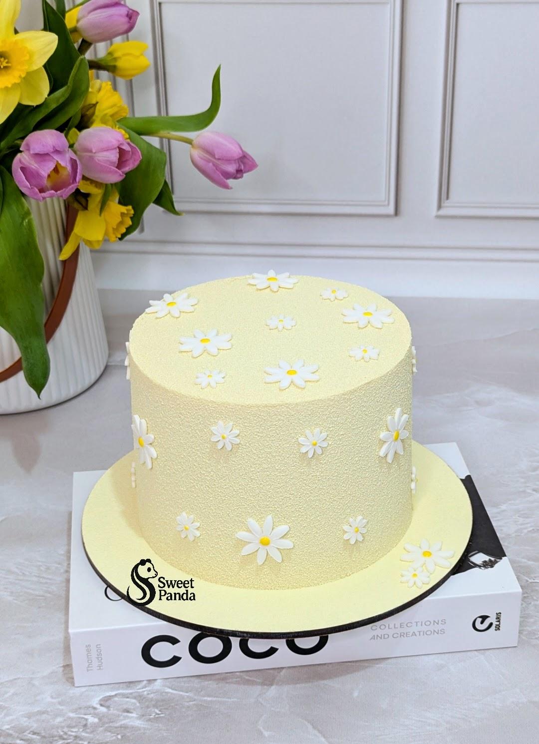 Two-tier pale yellow cake decorated with white and yellow daisy flowers on top and sides, displayed on white board box