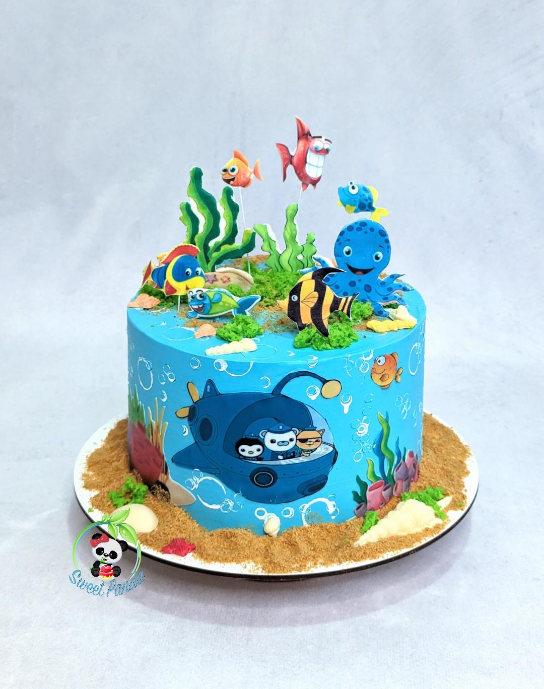 Single-tier ocean-themed cake with bright blue fondant, sand-textured base, fondant sea creatures including octopus